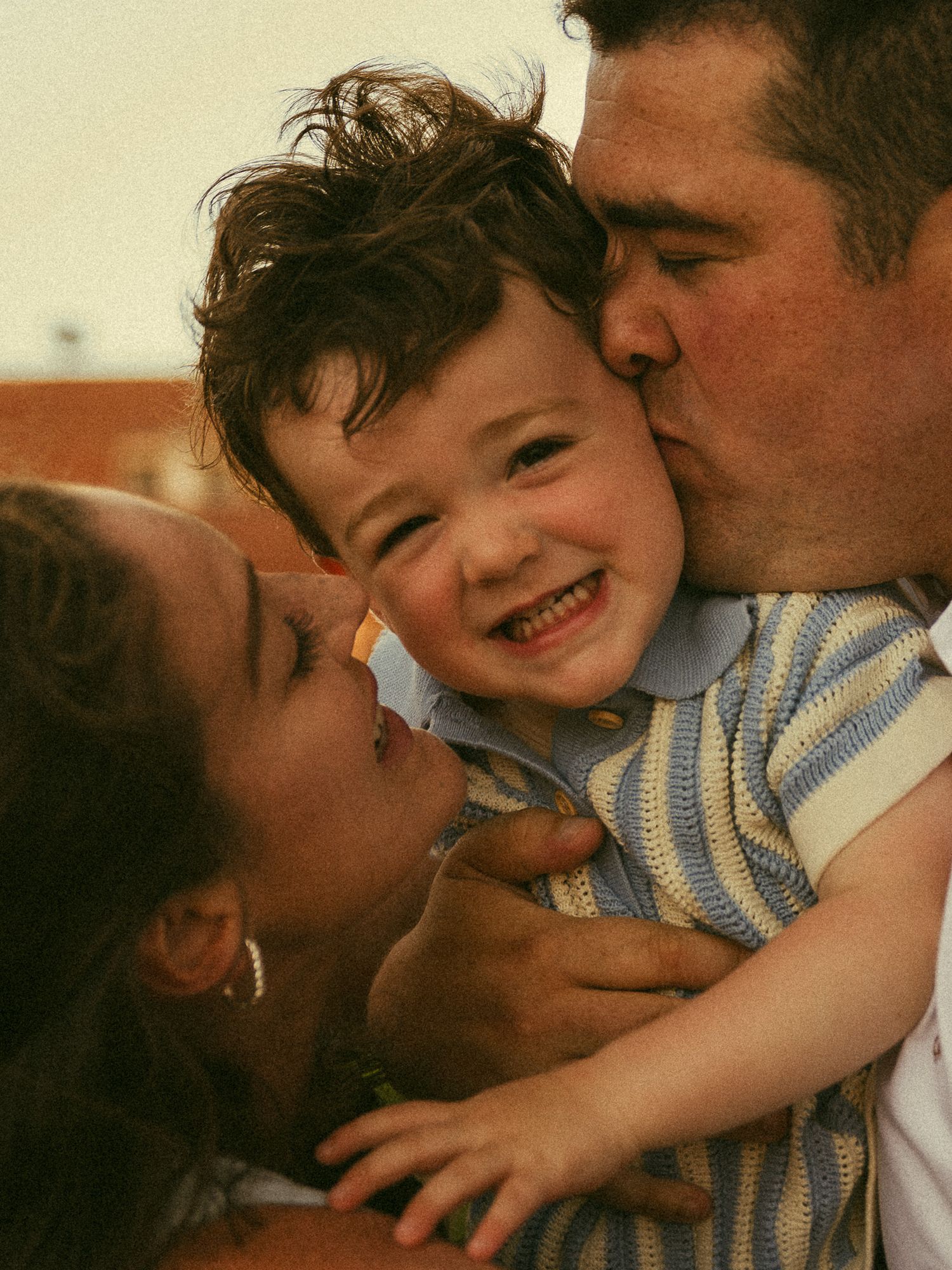 Happy toddler boy smiling while being kissed on both cheeks by his loving parents outdoors.