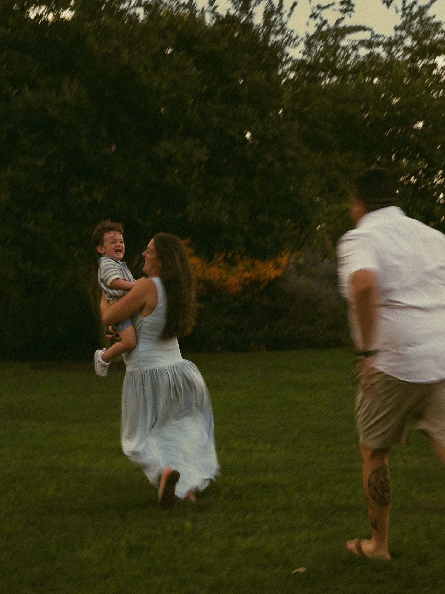 A woman in a white dress holds a baby while a man in a white shirt walks toward them on a green lawn.