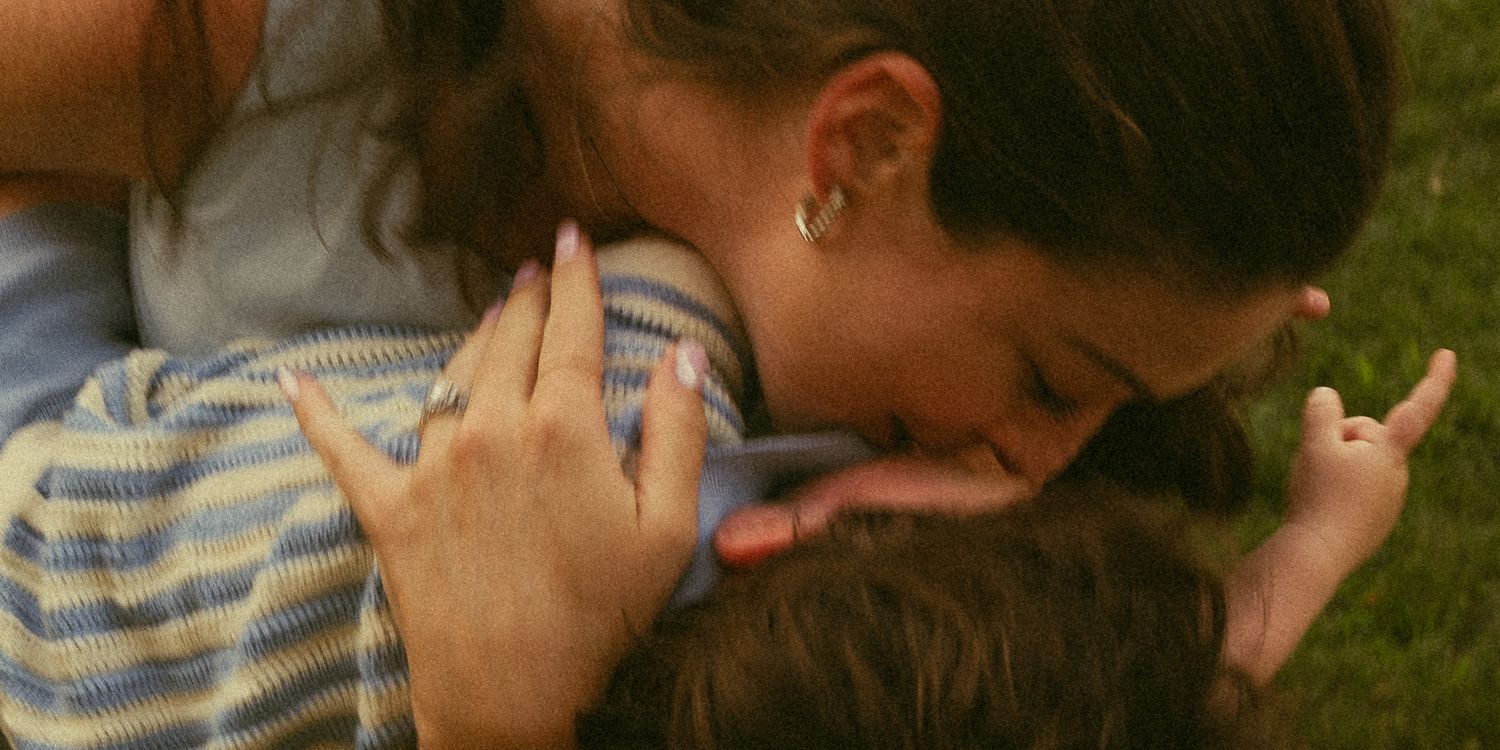 A woman tenderly kisses a child while lying on green grass outdoors.
