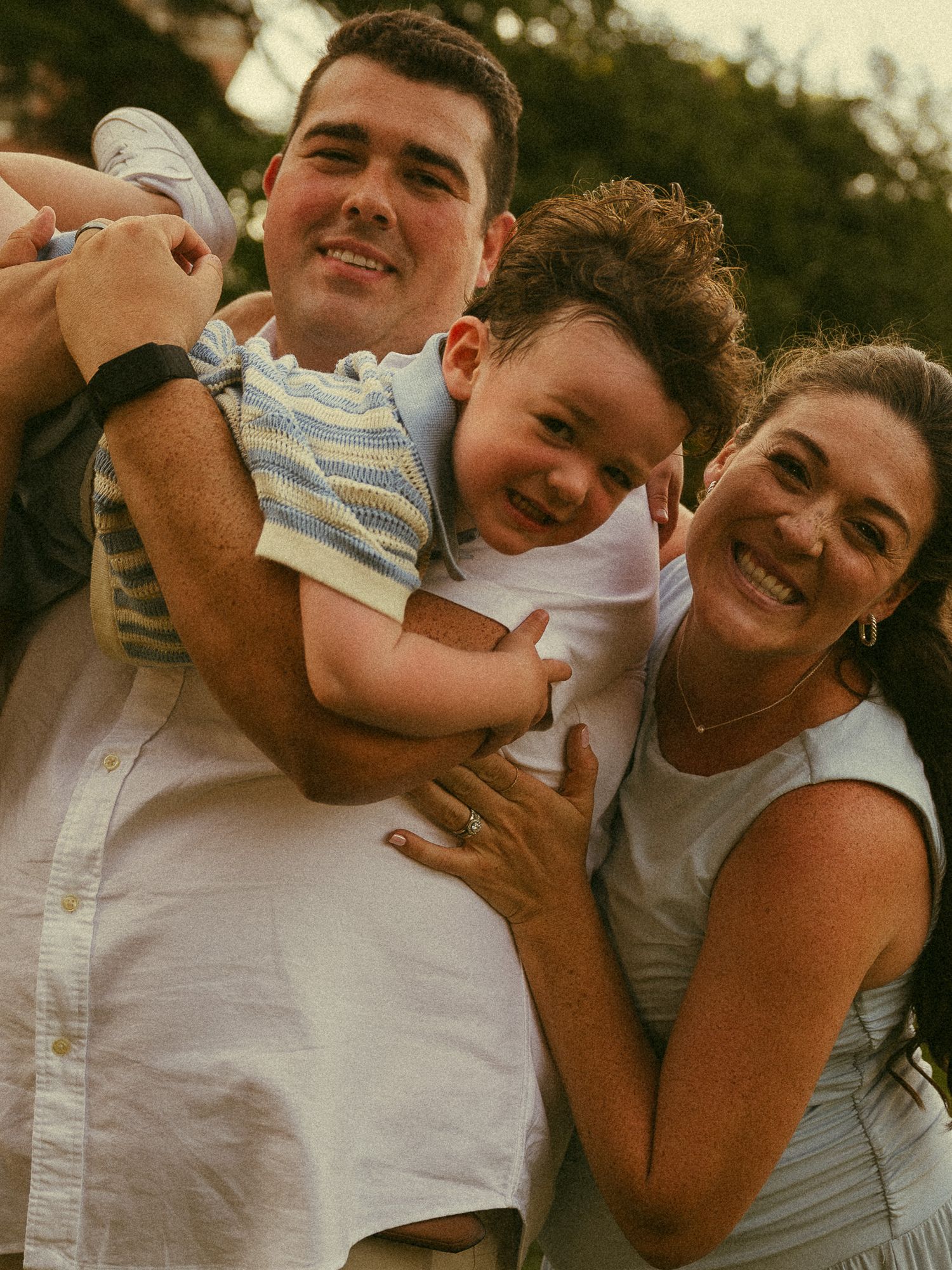 A happy family of three smiling outdoors, with a man and woman holding a laughing toddler in a blue striped shirt.