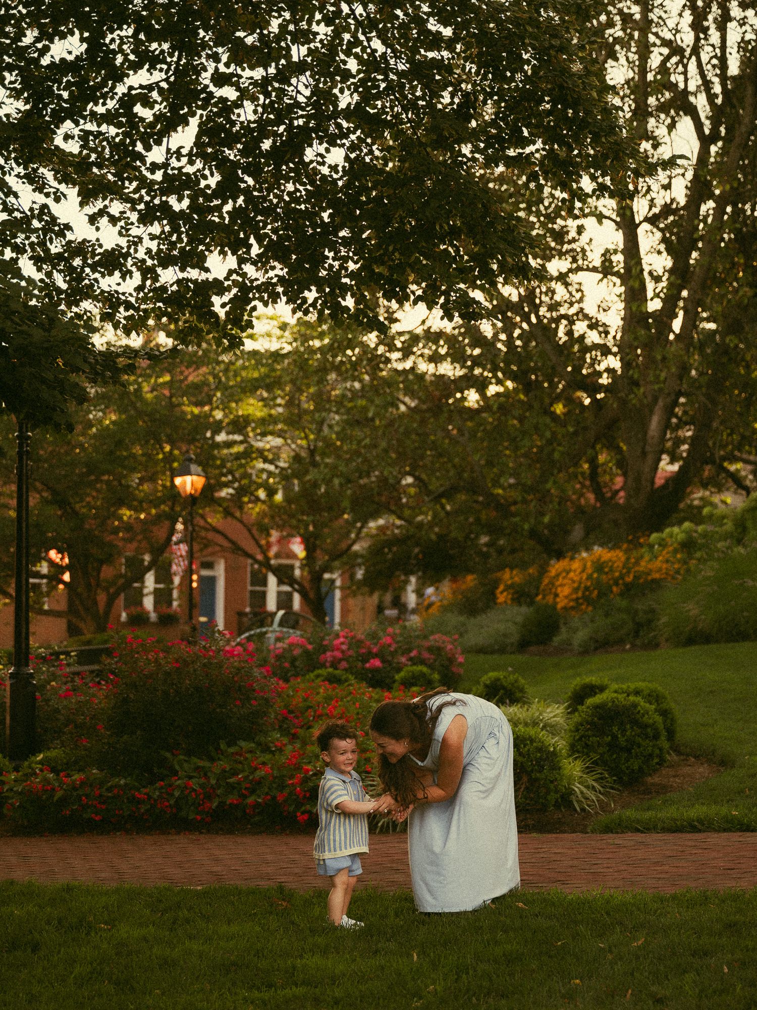 A mother bends down to hold hands with her toddler on a brick path surrounded by lush gardens at golden hour.
