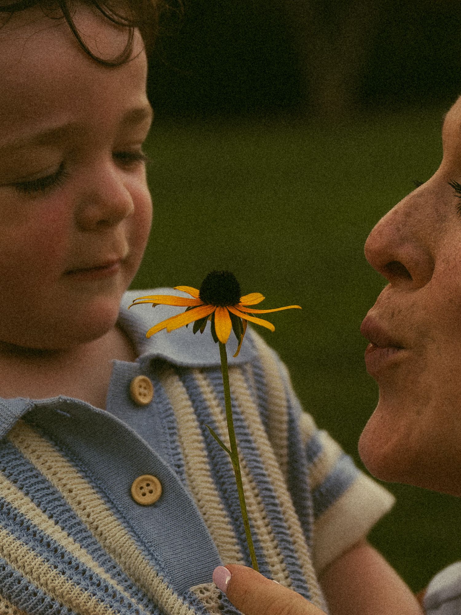 A young child and an adult share a tender moment smelling a yellow black-eyed Susan flower outdoors.