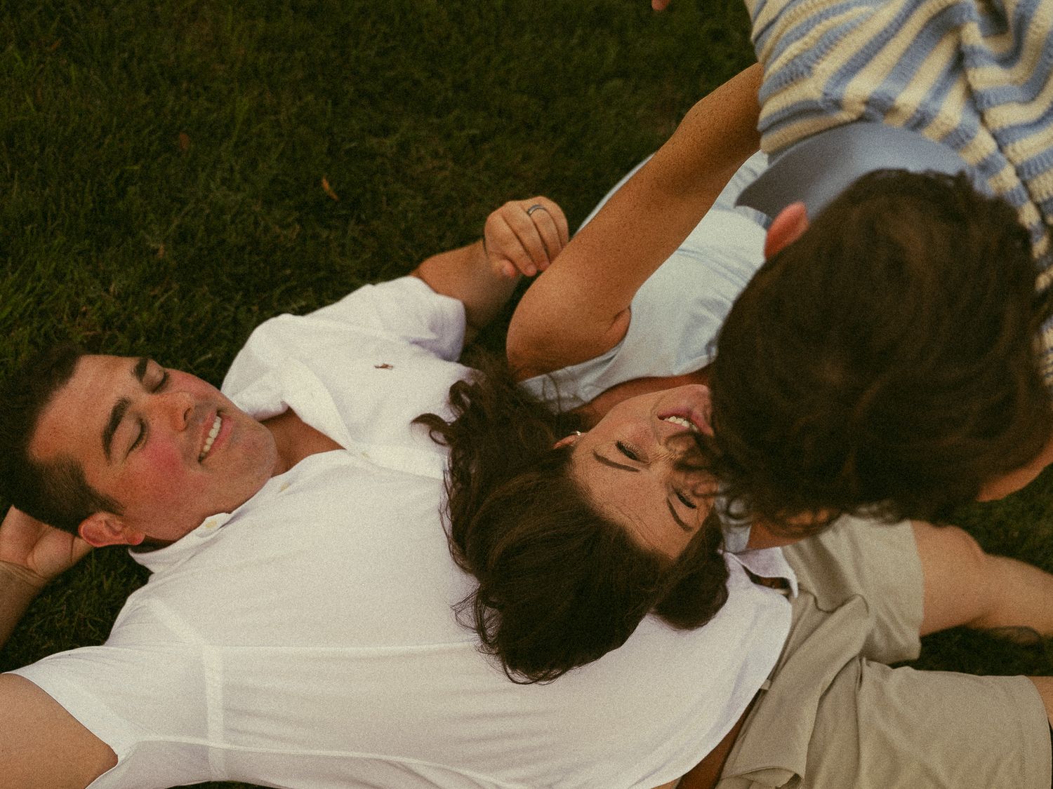 Three friends laughing and relaxing together on green grass outdoors on a sunny day.