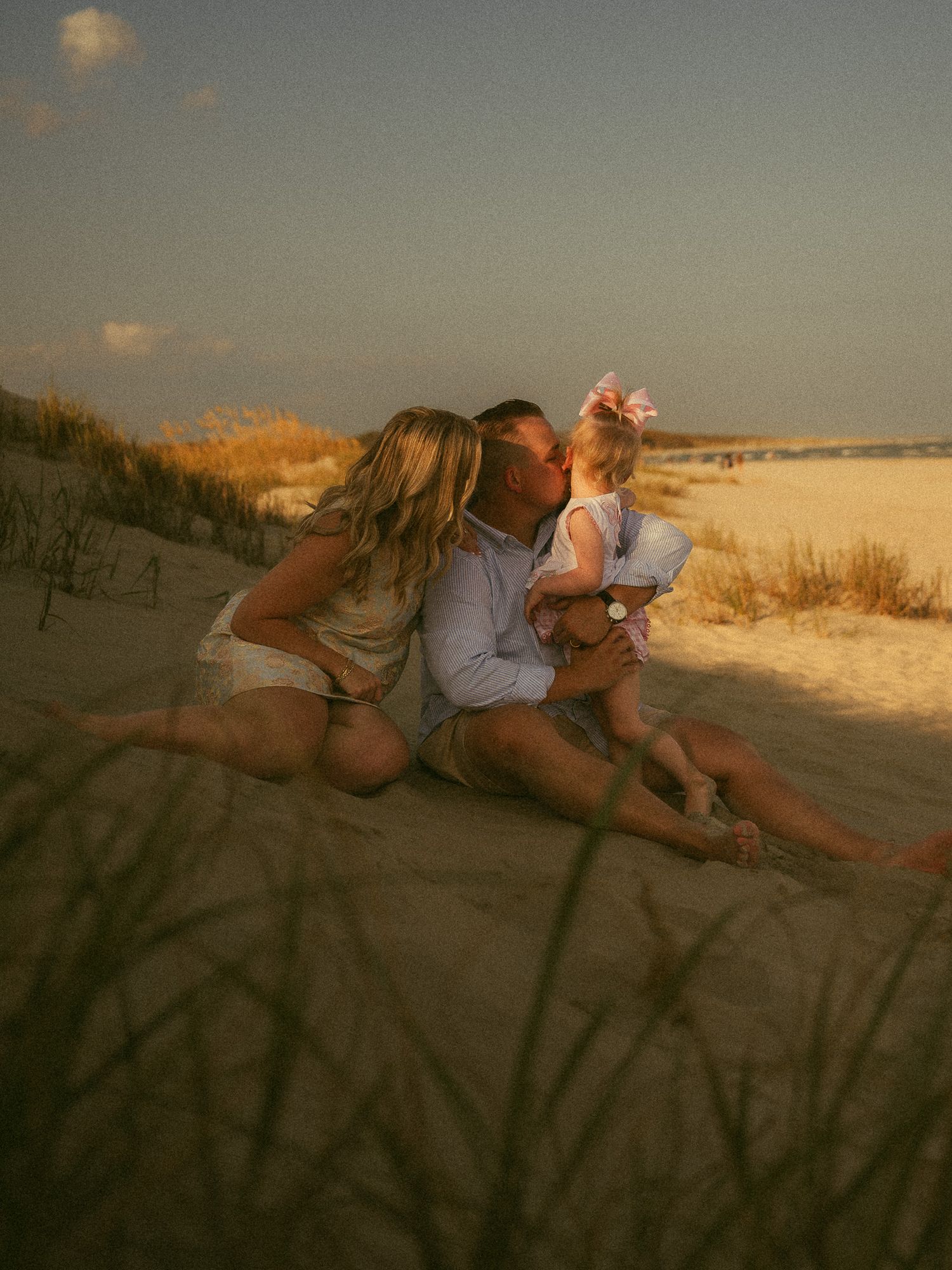 A couple shares a kiss on sandy beach dunes at sunset while holding their toddler daughter with a pink bow.