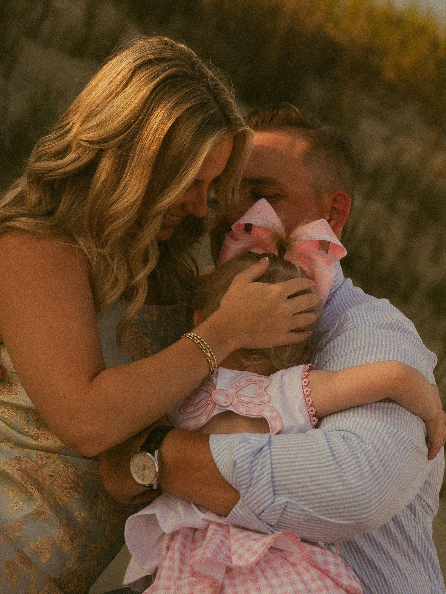 A couple shares a tender moment with their baby girl in a pink bow and dress during a golden hour family photo session.