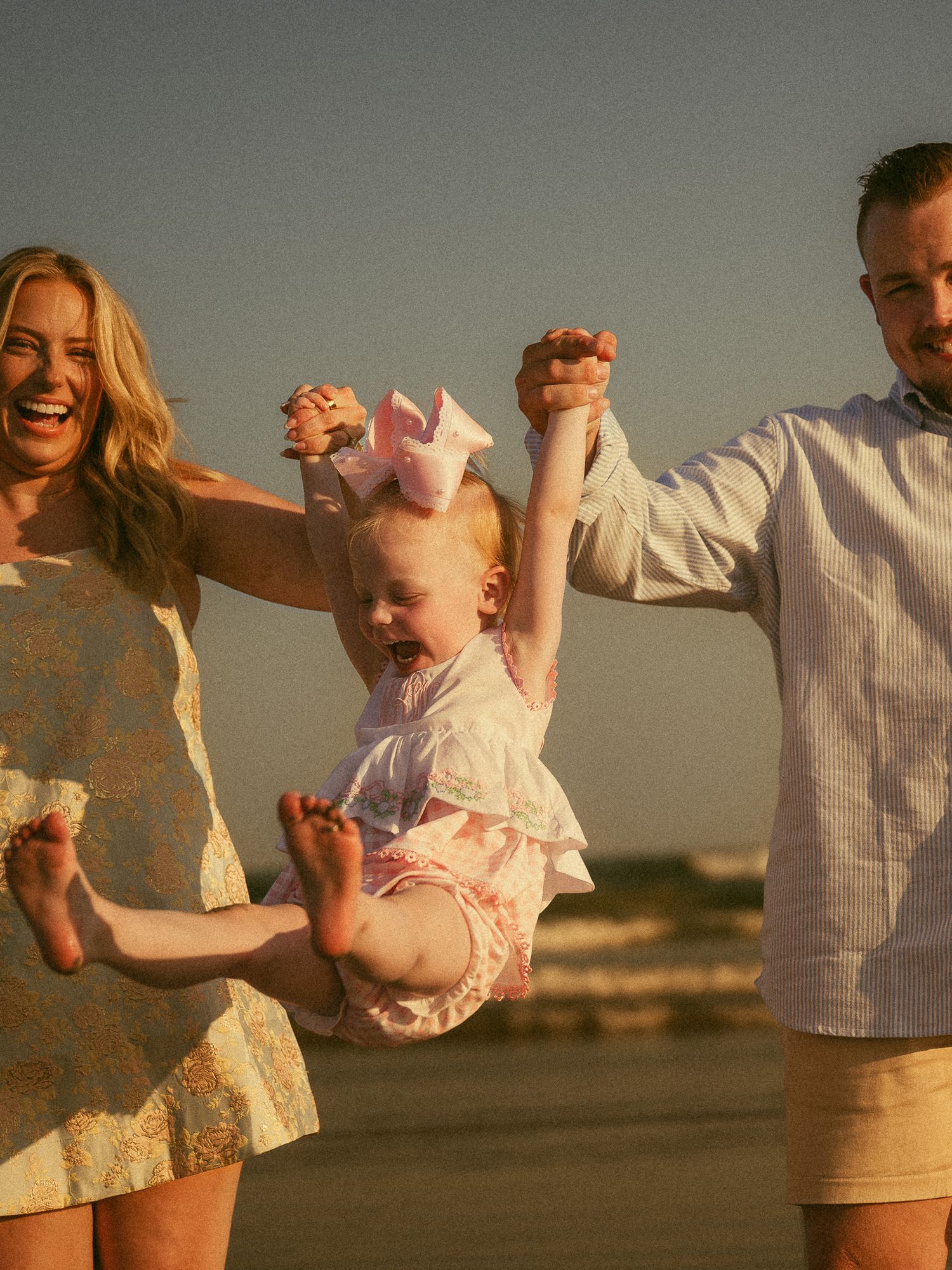 A joyful family swings their toddler girl on a sunny beach, with mom in floral dress and dad in blue shirt.