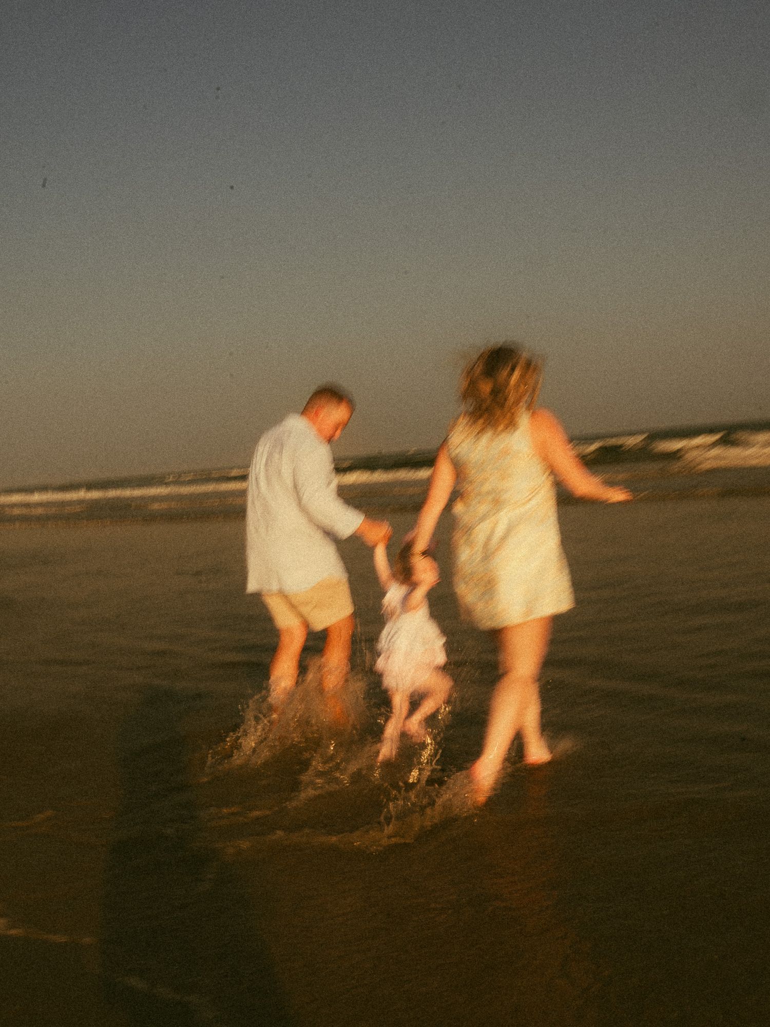 A blurry photo of a family with a toddler playing in ocean waves at sunset on a sandy beach.