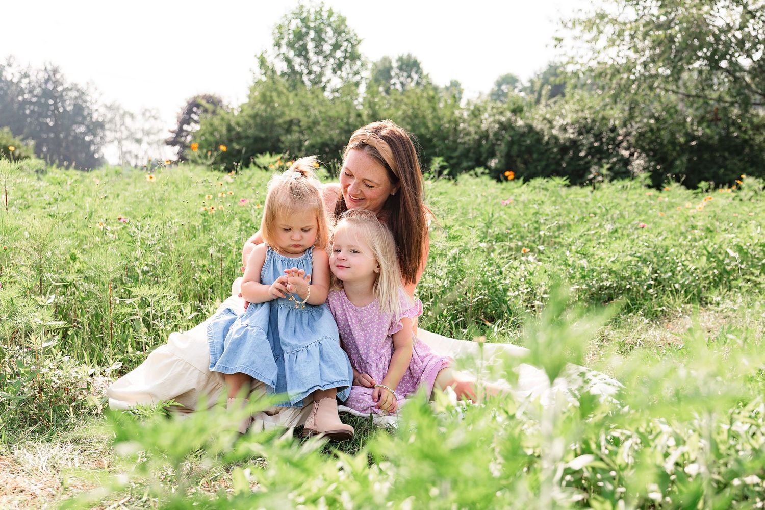 A mother sits in a lush green meadow with her two young daughters, sharing a tender moment outdoors.
