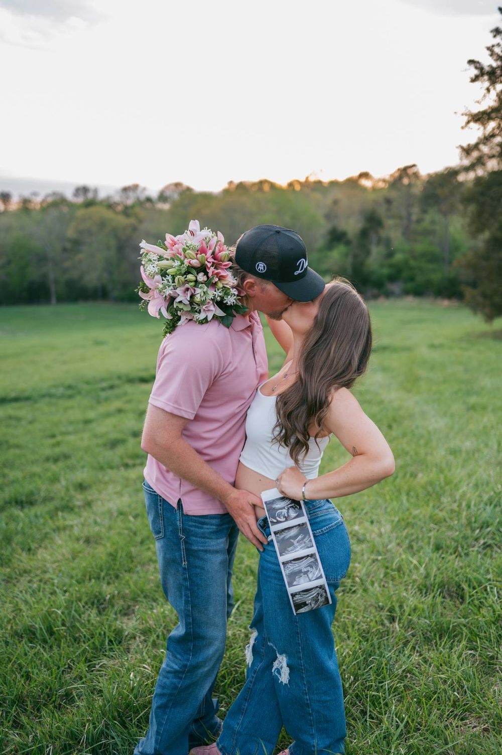 Couple kissing in green field at sunset, woman holding ultrasound photos and man holding pink flowers Asheboro NC