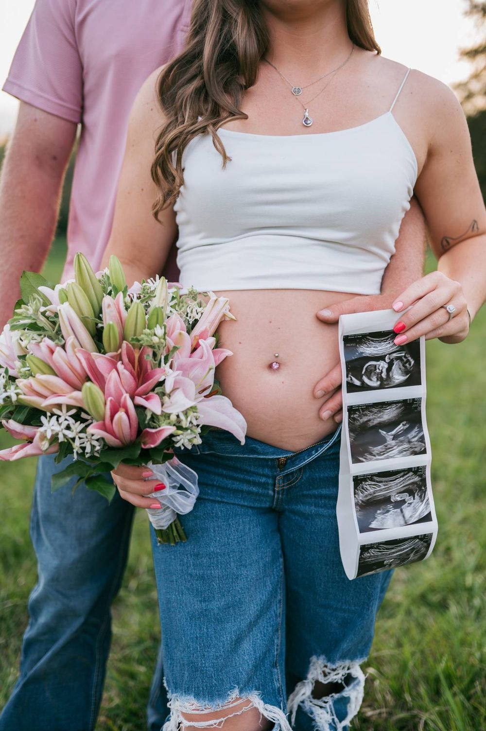 Close-up of pregnant woman's bare belly holding ultrasound photos and pink lily bouquet in field in North Carolina