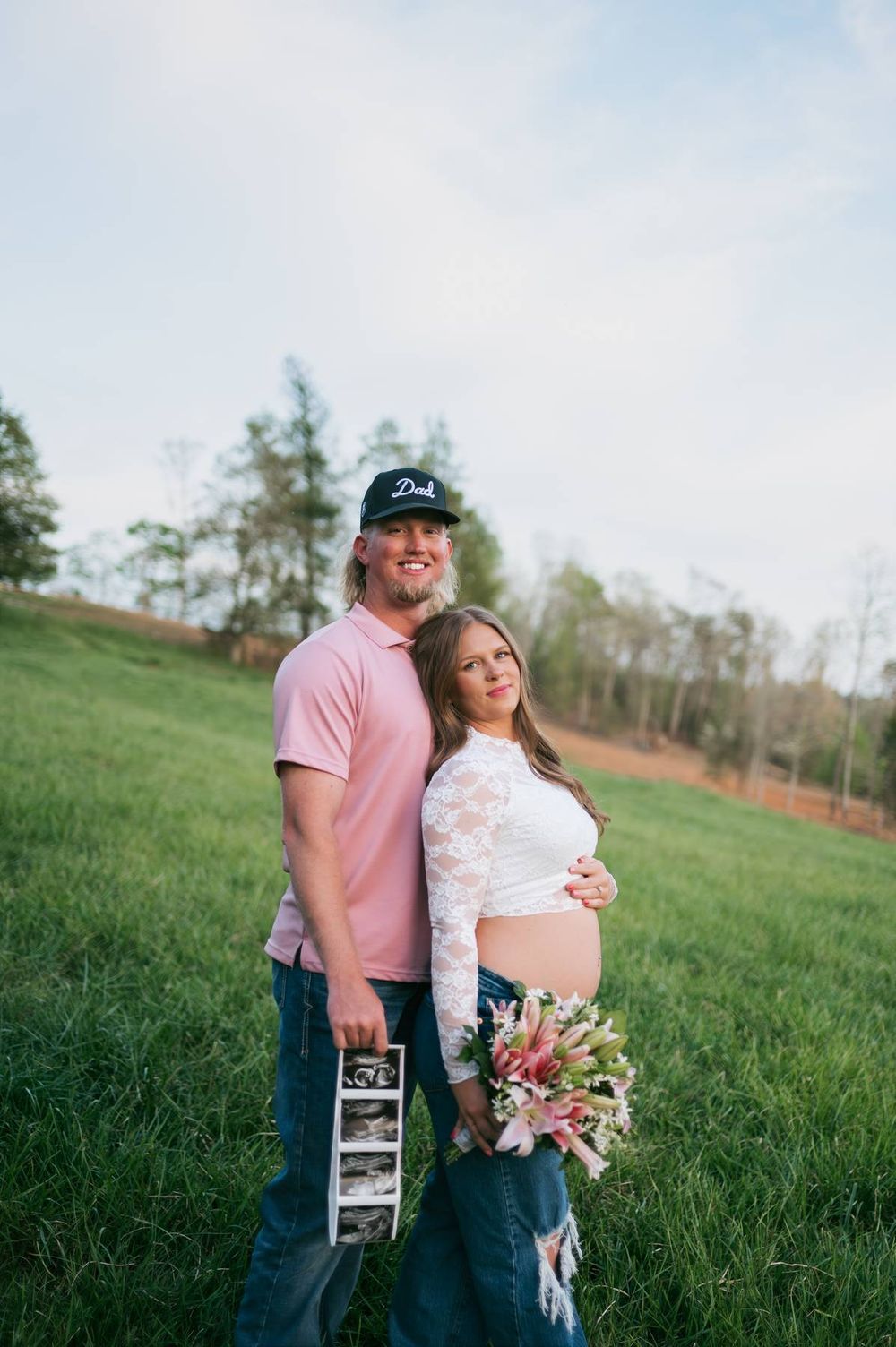Smiling couple in green field, man in pink shirt and Dad cap, pregnant woman in lace crop top North Carolina