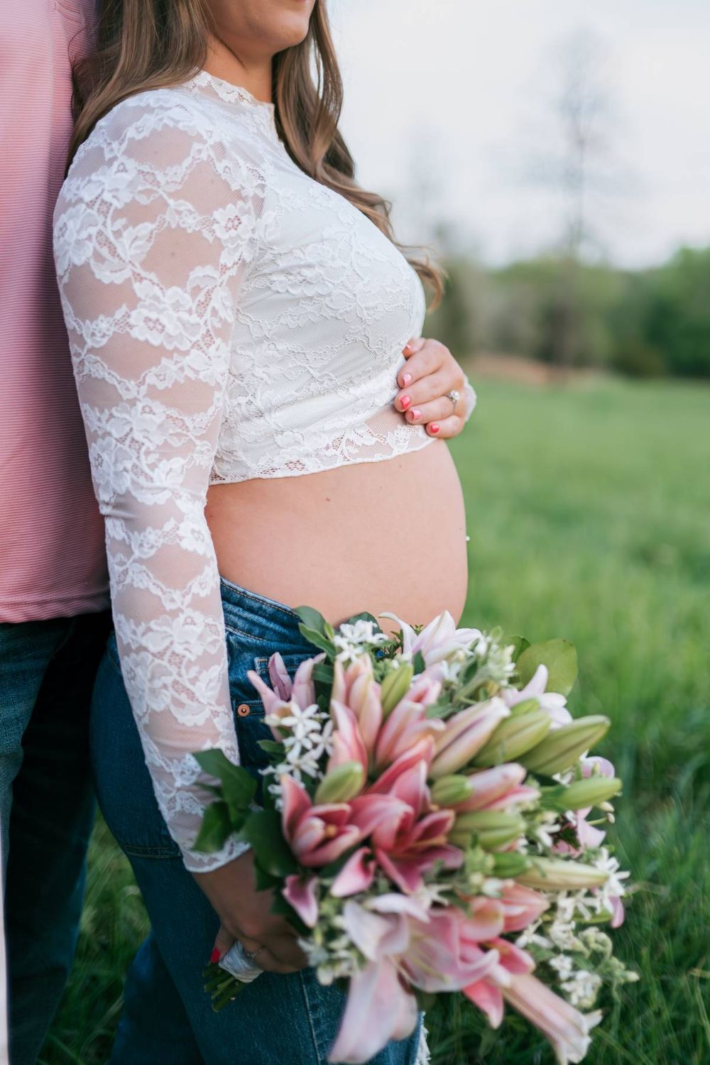 Close-up of pregnant belly in lace crop top and jeans with pink lily bouquet in green field Lexington NC