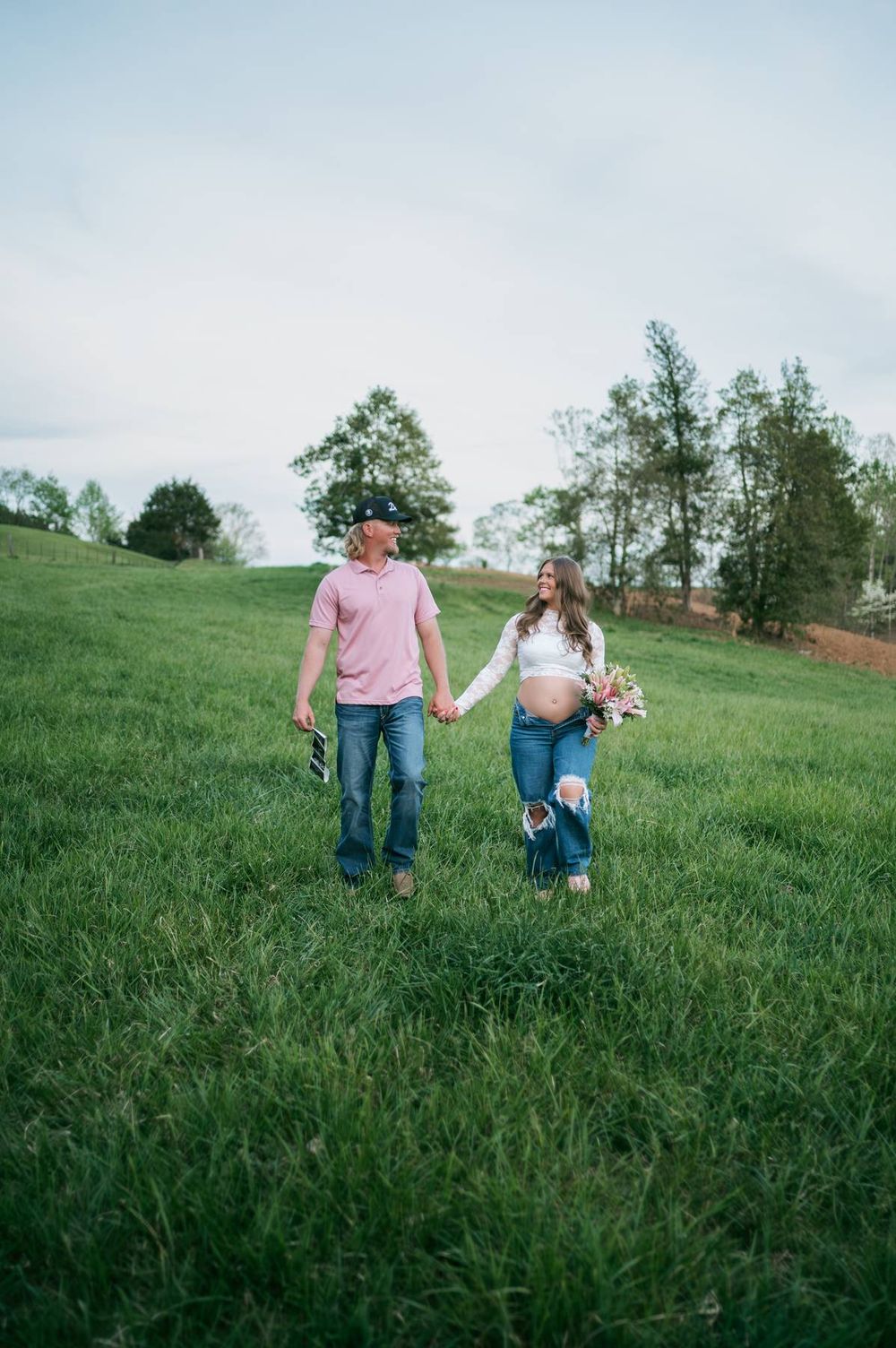 Couple holding hands walking through lush green field, pregnant woman in lace top carrying flowers High Point NC