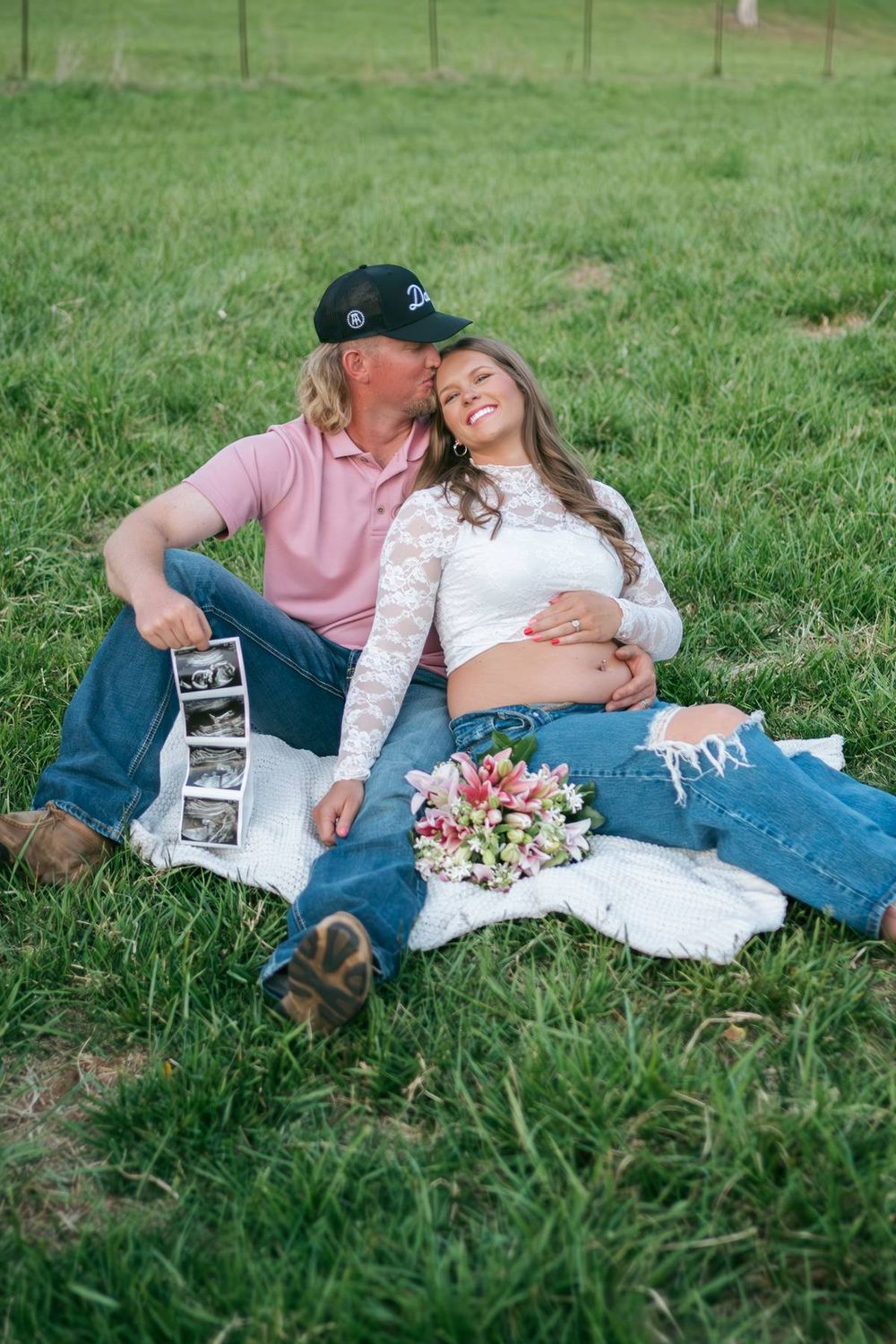 Couple sitting on grass blanket during outdoor maternity shoot, holding ultrasound photos and pink flowers Greensboro NC