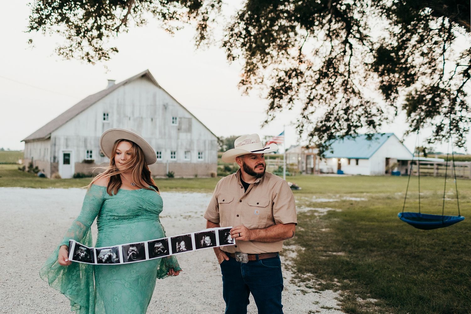 Pregnant couple in cowboy hats holding ultrasound strip photos on a farm with a white barn in the background.