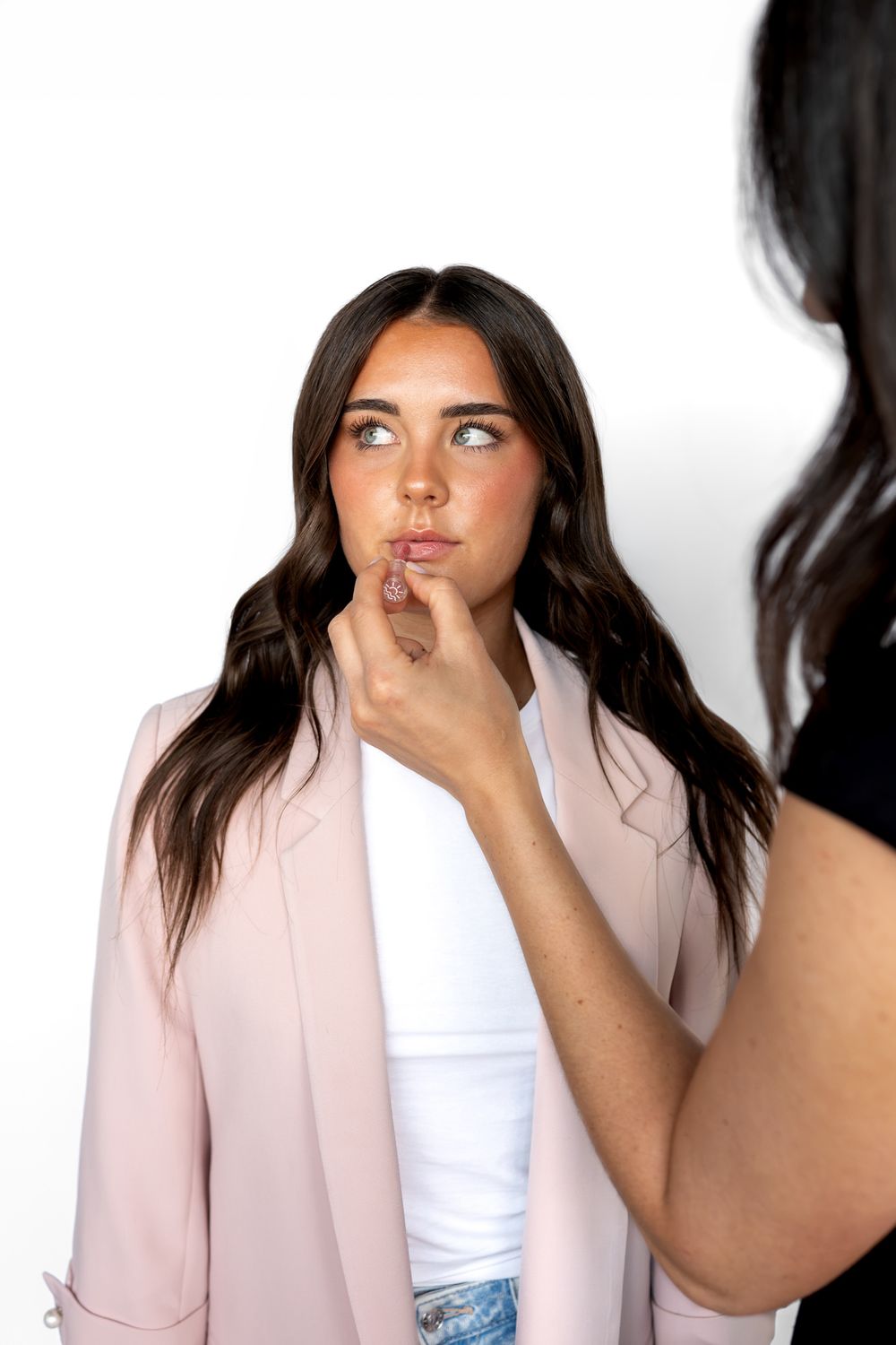 Makeup artist applying lip product to brunette woman in pink blazer against white background.