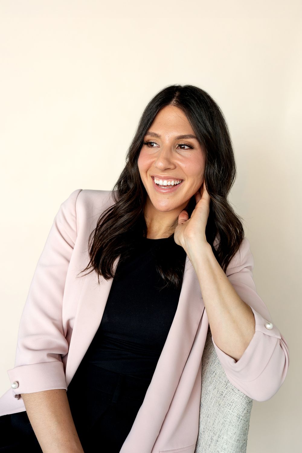 Smiling woman with dark hair wearing a pink blazer over black top, posing confidently against a cream background.
