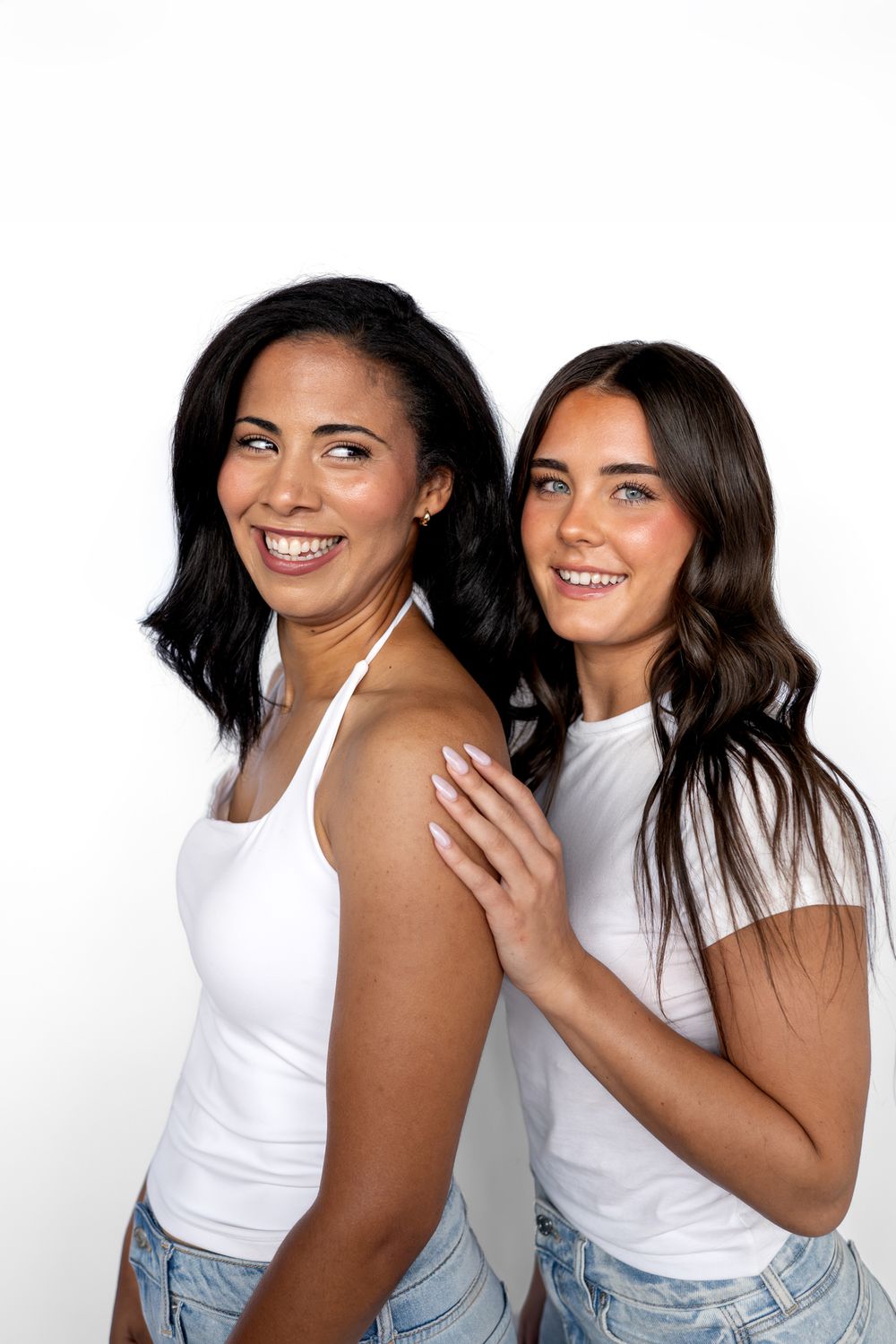 Two smiling women in white tops and denim shorts pose together against a white background in a bright studio setting.