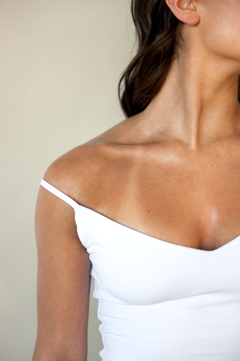 Close-up of a woman's neck, shoulder, and décolletage in a white off-shoulder top against a neutral background.