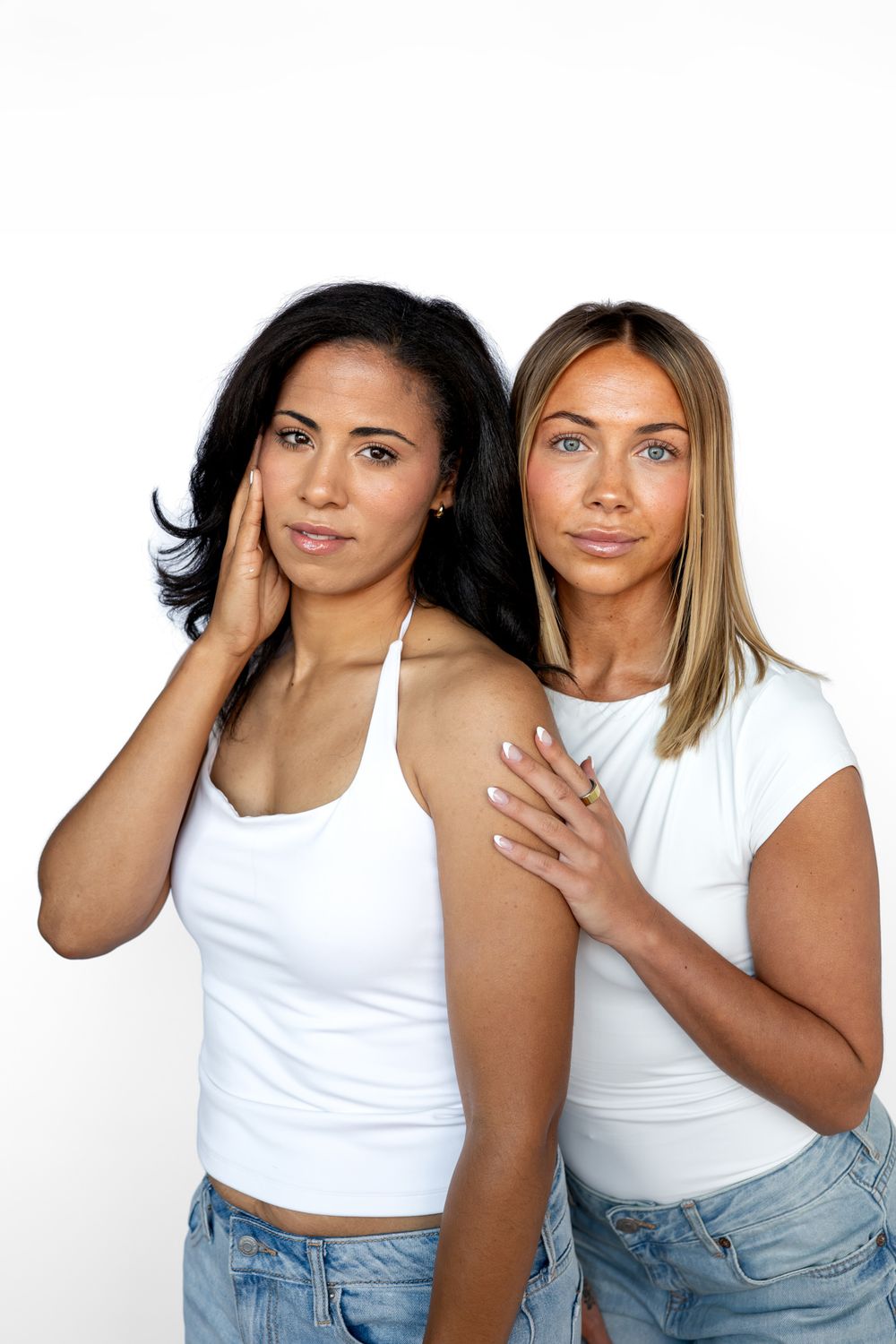 Two women in white tops and jeans pose together against a white background.
