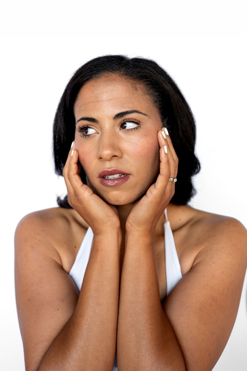 Worried young woman in white top holding face in hands with wide eyes against white background.