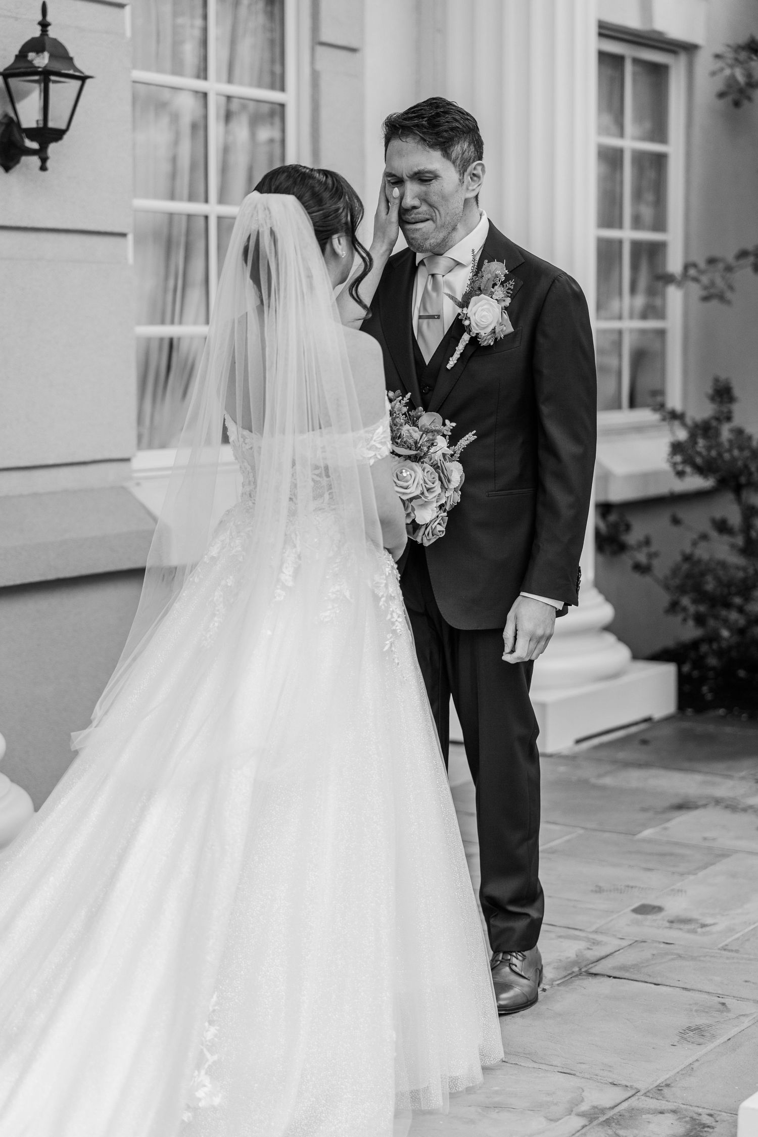 Black and white photo of emotional groom wiping tears as bride in lace gown and veil faces him outside elegant venue.