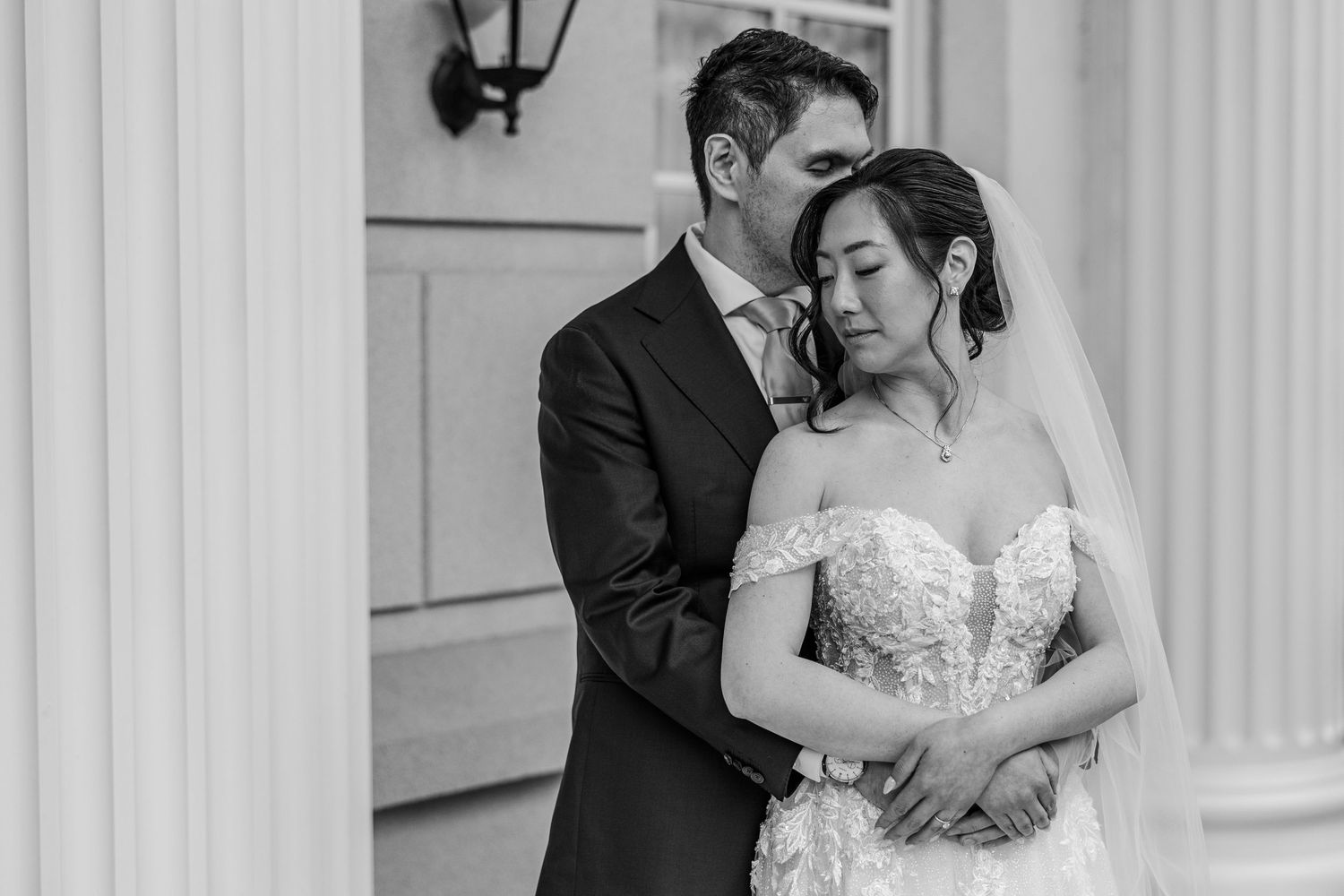 Black and white wedding photo of bride and groom embracing by white columns.