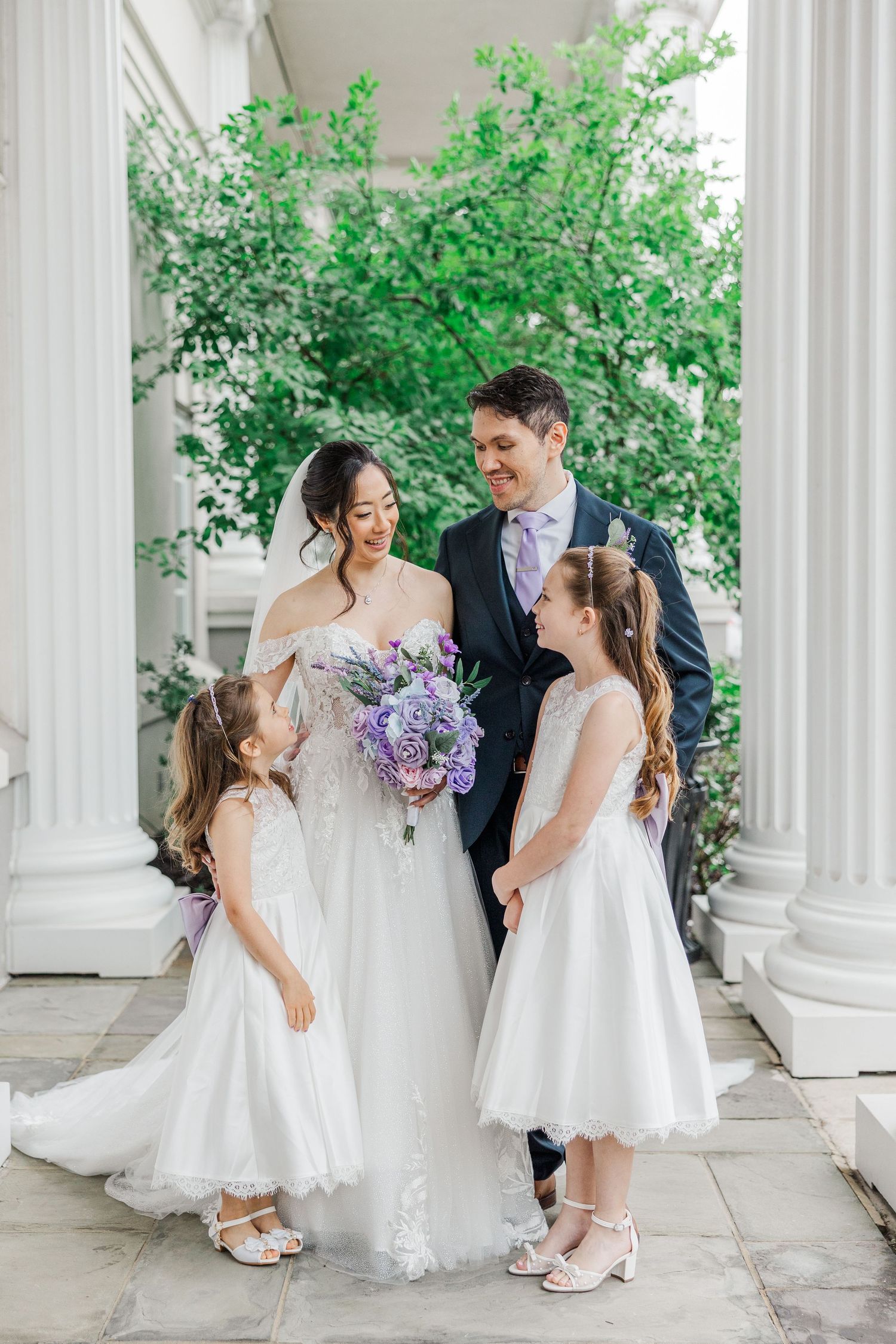 Bride and groom with two flower girls in white dresses, posing by white columns with lush green trees.