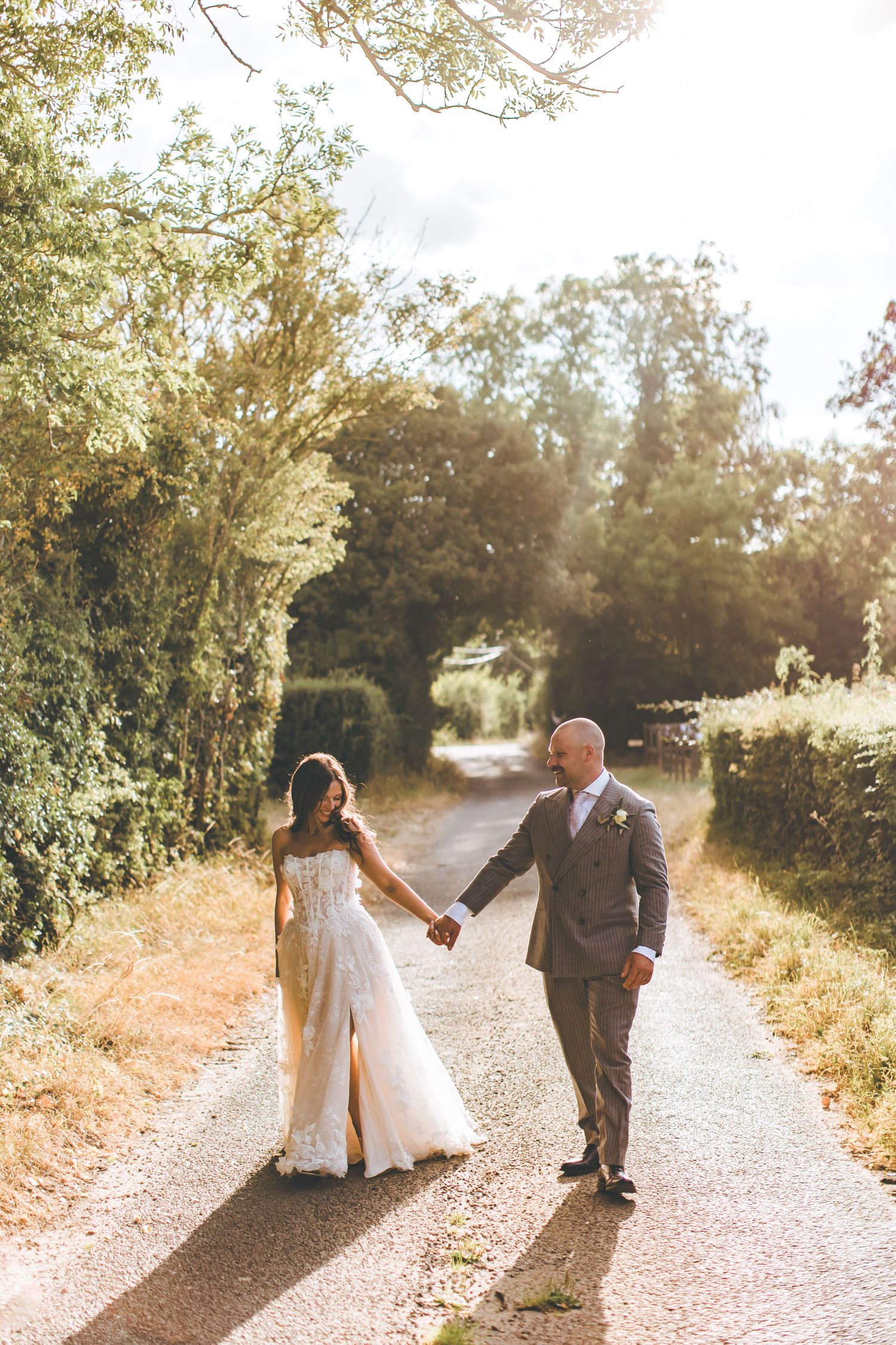 Bride and groom holding hands walking down a sunlit country lane lined with lush green trees on their wedding day.