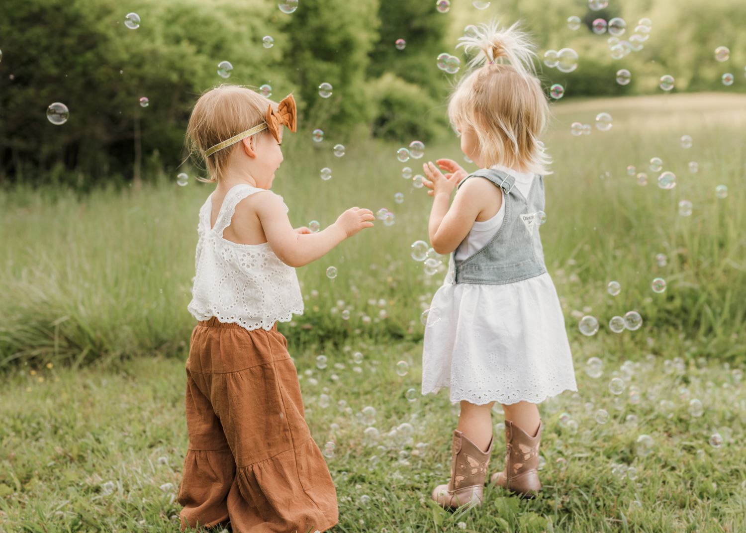 Two toddler girls playing with bubbles in a green meadow, wearing cute summer outfits.