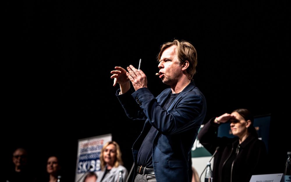 A conductor leads an orchestra on stage, holding a baton while performers stand behind him in a dimly lit venue.