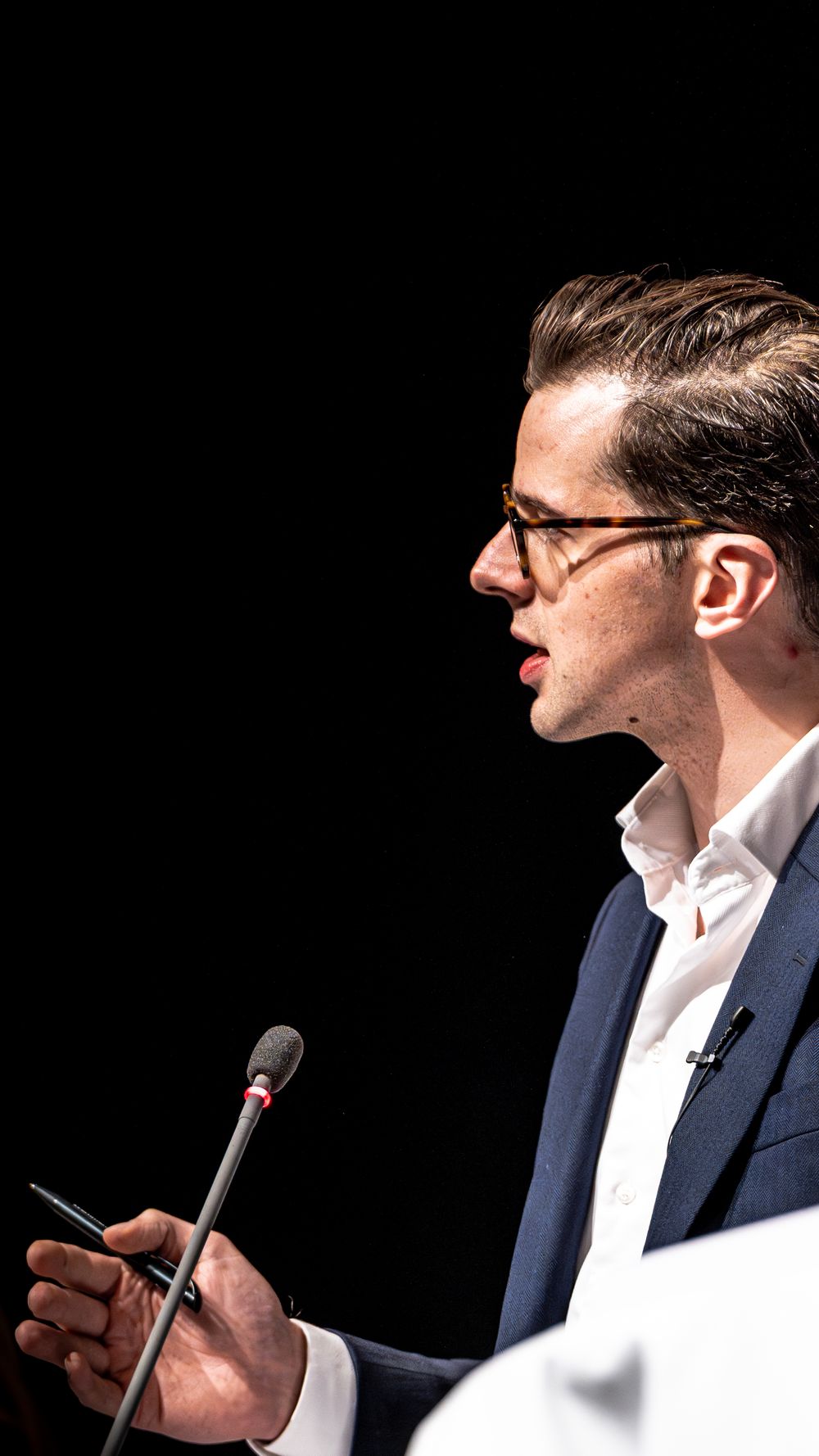 Young man in glasses and navy blazer speaking at podium with microphone against dark background.