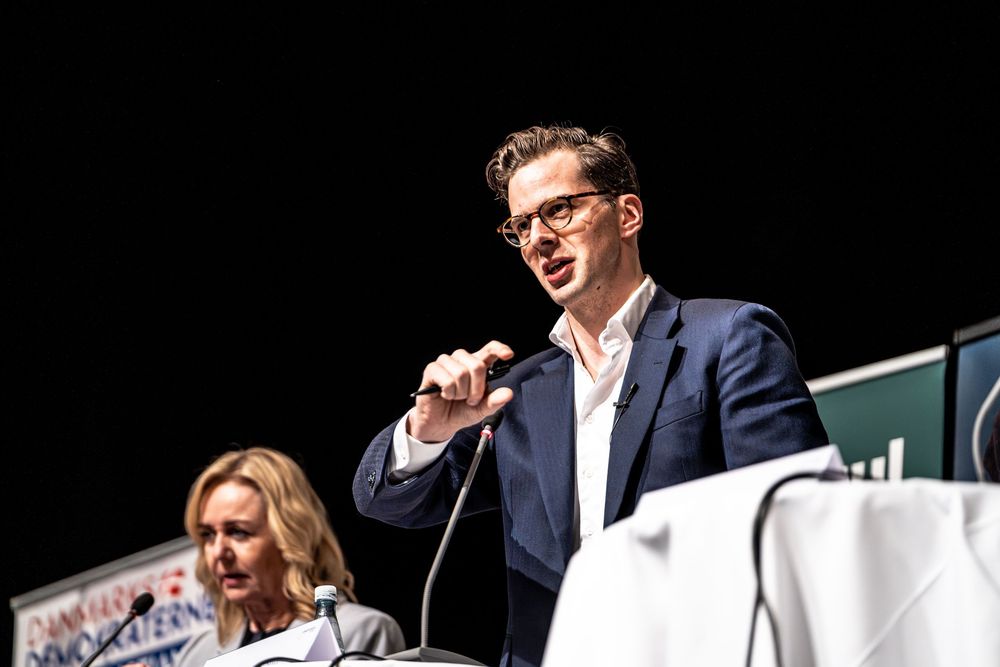 Young man in blue blazer speaks at podium with microphone at formal event, woman seated behind him.