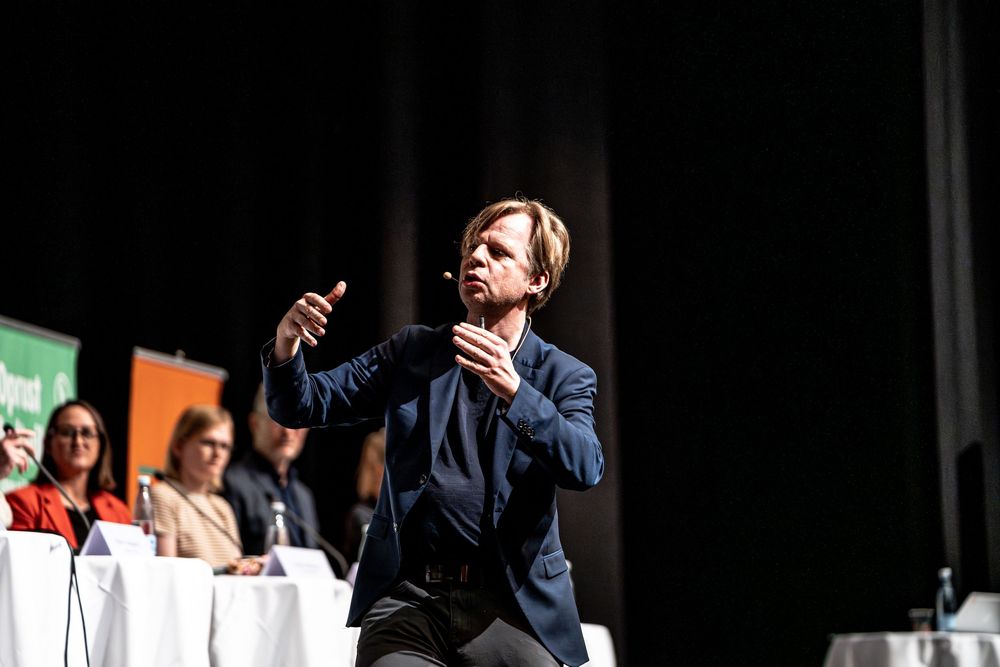 A speaker in a navy blazer gestures expressively on stage during a panel discussion event.