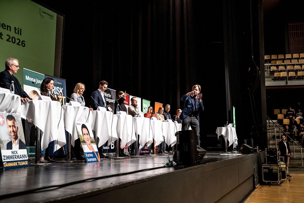Panel debate on stage with multiple participants behind white podiums at a political event in 2026.