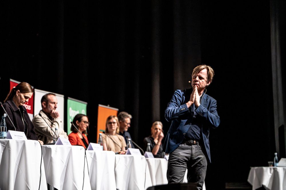 Panel discussion on stage with seated speakers behind white-draped tables and a standing presenter with hands clasped.