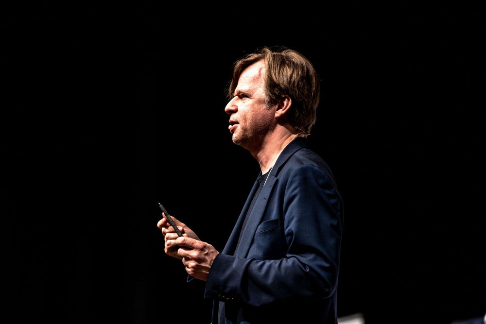 Man in navy blazer speaking on stage, holding a phone, illuminated against a dark background.