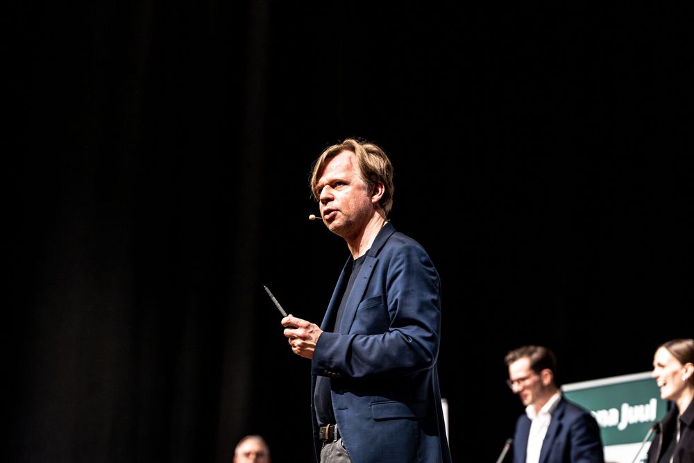 Man in blue blazer speaking on stage, holding a microphone at a professional conference event.
