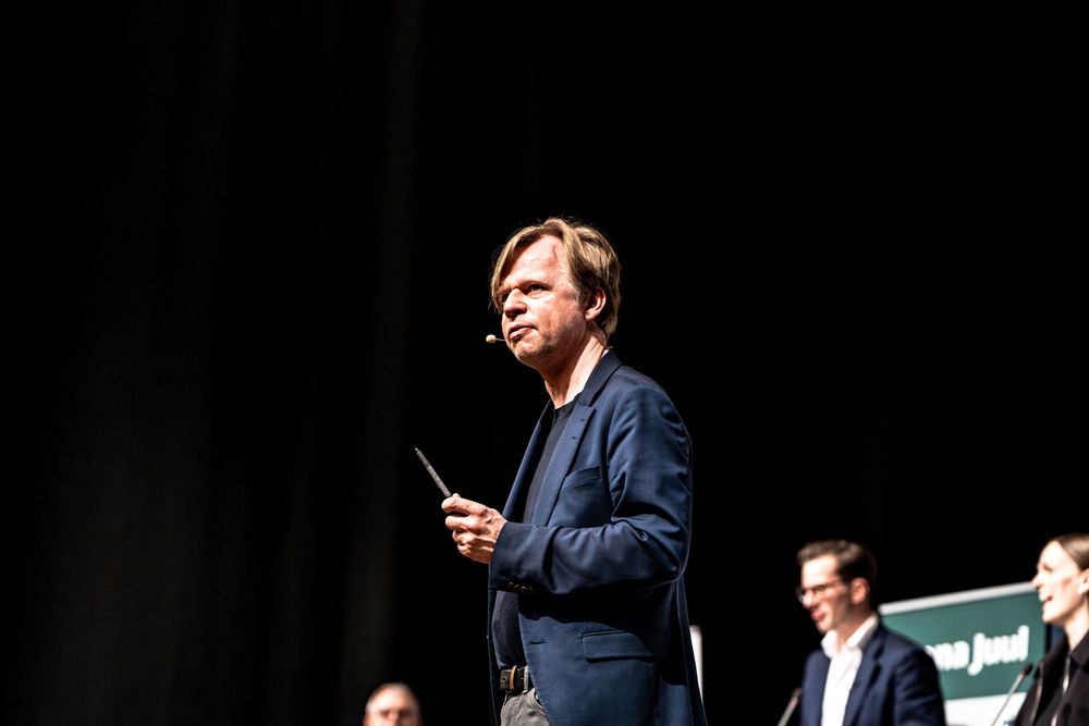 Man in navy blazer speaking on stage with headset microphone, holding a device during a conference presentation.