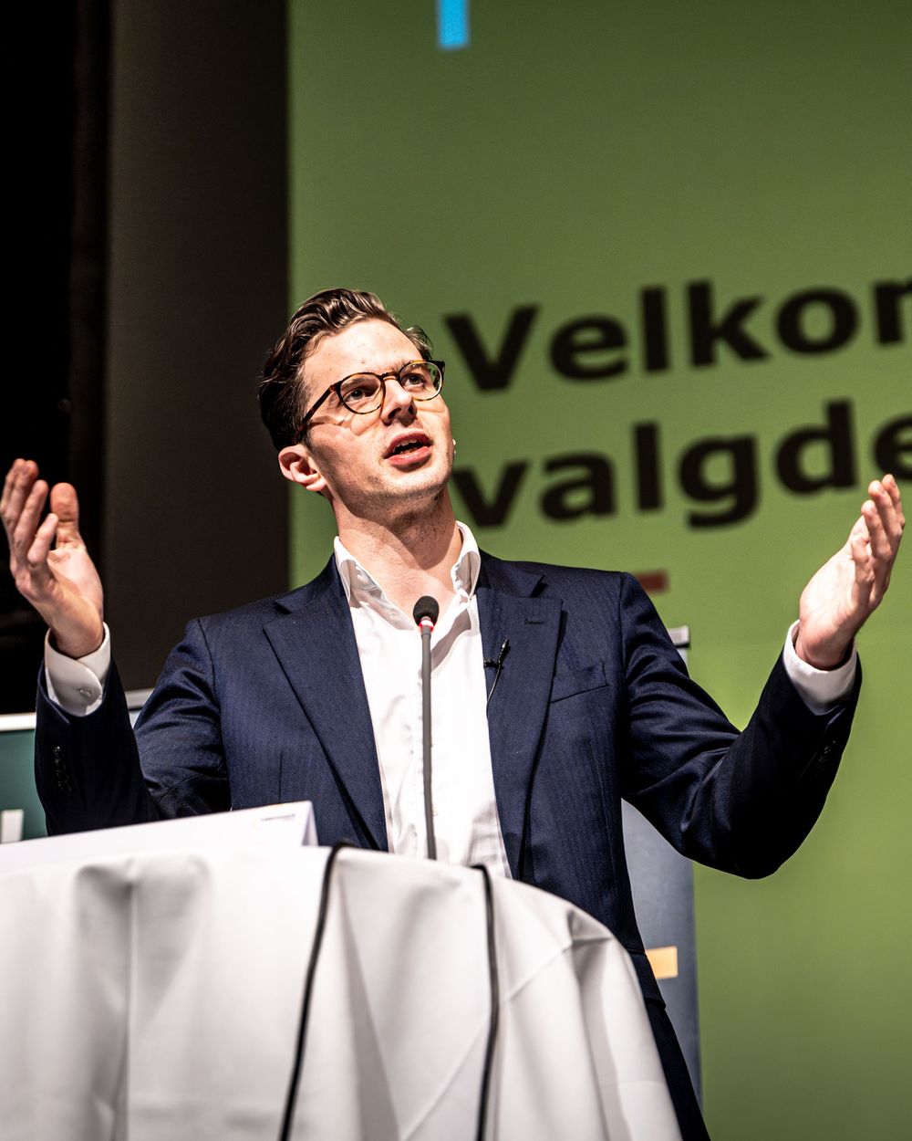 Man in navy suit gestures at podium during Norwegian political event with green backdrop reading Velkommen.