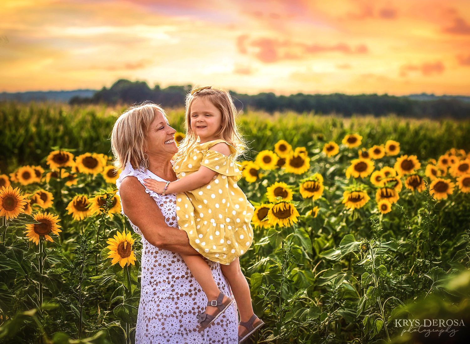 Woman holding young girl in yellow dress among sunflowers at golden sunset, smiling joyfully in a vibrant field.
