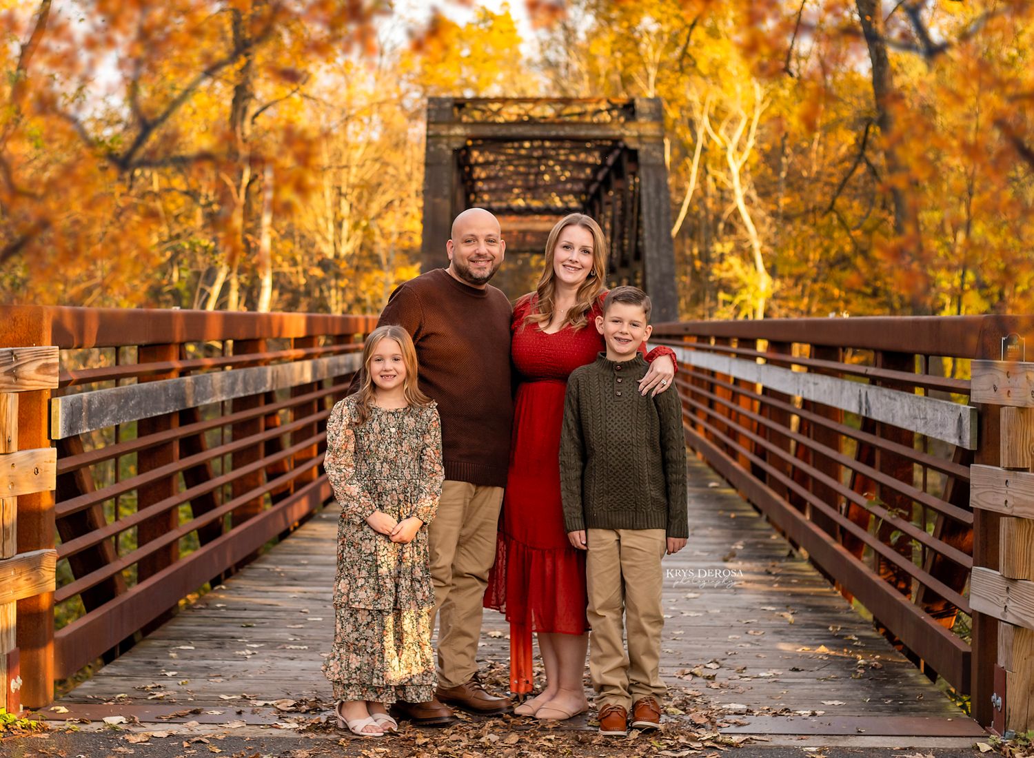 Family of four posing on a wooden bridge surrounded by vibrant autumn foliage for a fall portrait.
