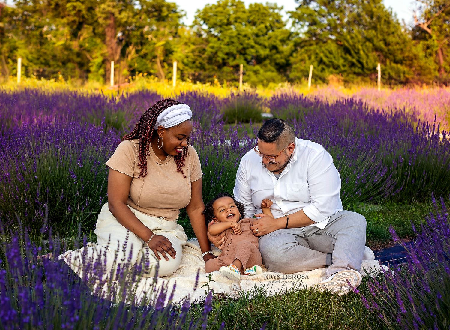 Family sitting in lavender field at golden hour, parents smiling at newborn baby on blanket.