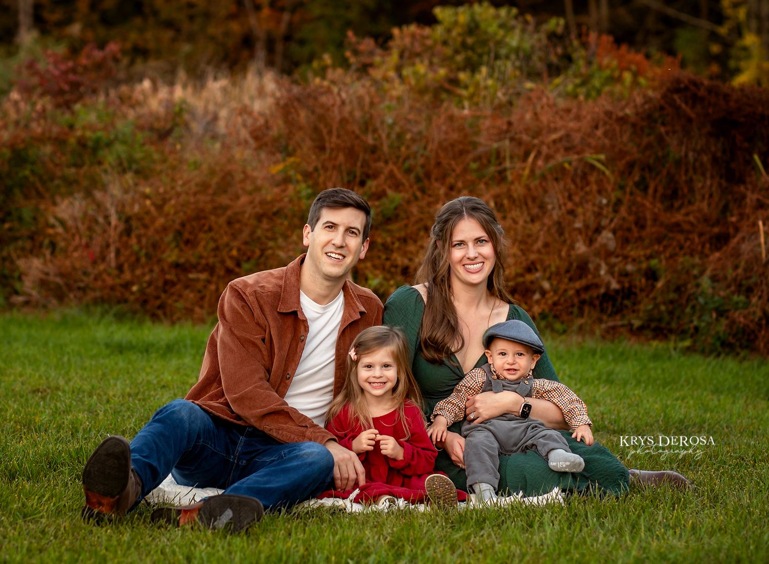 Family of four smiling outdoors in autumn, parents seated on grass with young daughter and baby son in colorful fall foliage.