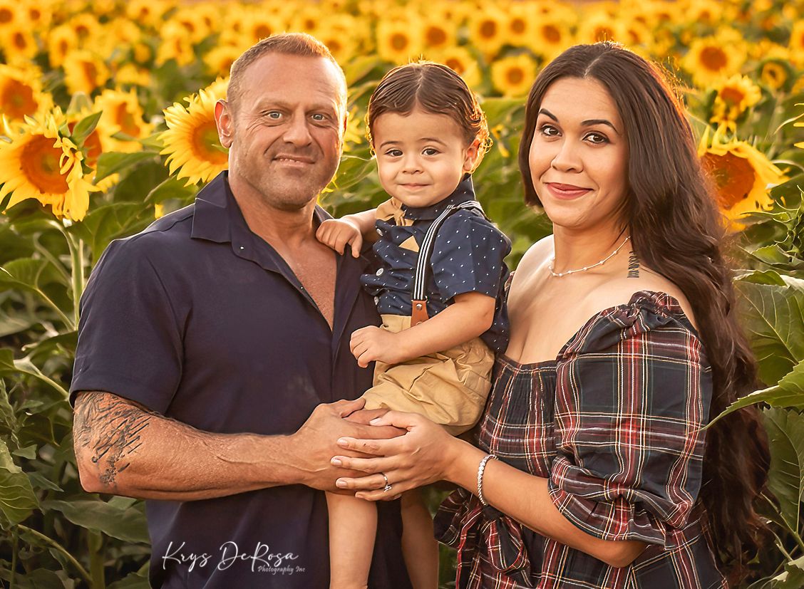 Family of three posing in a vibrant sunflower field, parents holding toddler, warm golden hour lighting.