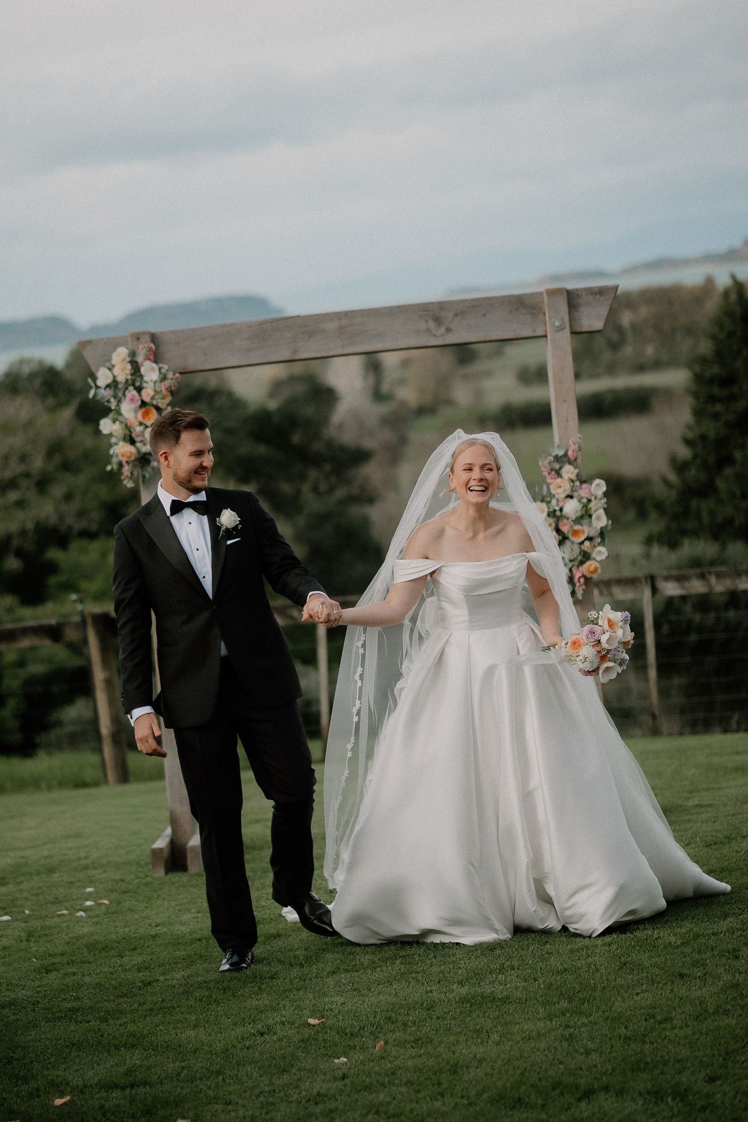 Black and white photo of a smiling bride in off-shoulder gown embracing groom in tuxedo, veil flowing in the wind.
