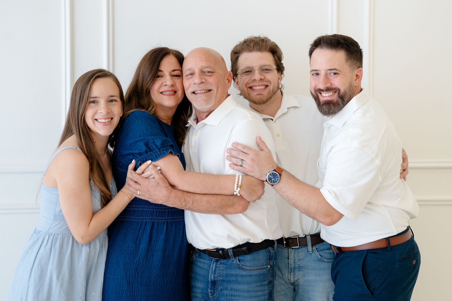 A joyful family of five poses together smiling in a bright white studio, wearing casual blue and white outfits.