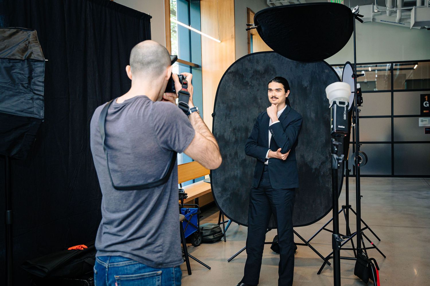Photographer taking professional headshot of man in suit against dark backdrop with studio lighting equipment.