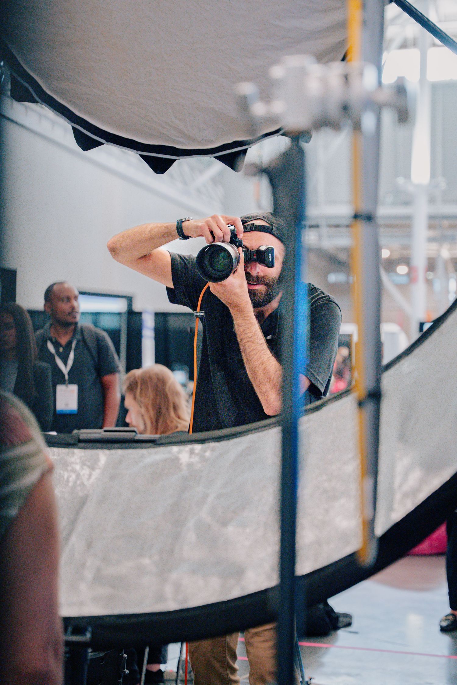 Bearded male photographer holding a DSLR camera at a photography expo, reflected in a curved reflector panel.