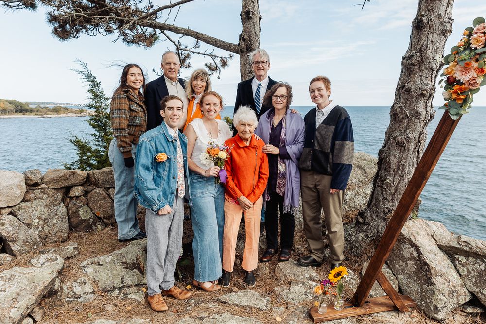 Family group photo outdoors near rocky shoreline with pine trees, posing together at a coastal wedding ceremony.